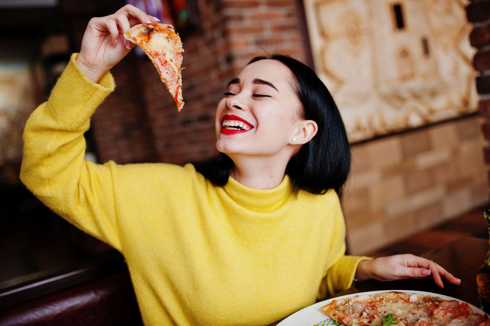 Funny brunette girl in yellow sweater eating pizza at restaurant.